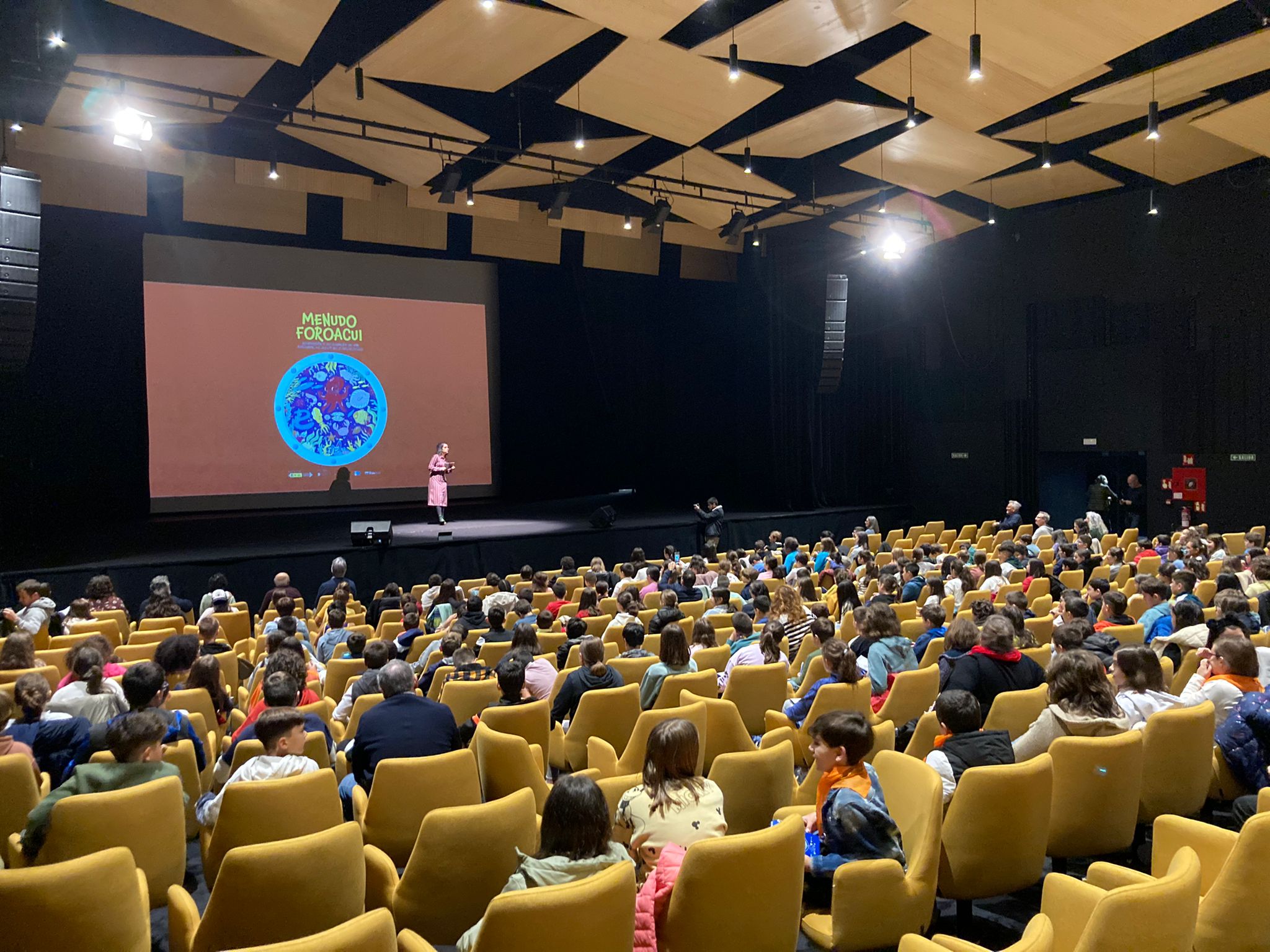 Apertura de MenudoForoAcui. Auditorio cheo de crianzas, coa presentadora no escenario e a imaxe do proxecto na pantalla do fondo.